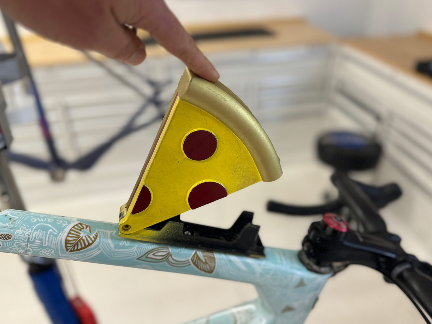 A hand pointing at a playful pizza slice bike accessory attached to a bicycle frame in a workshop setting.