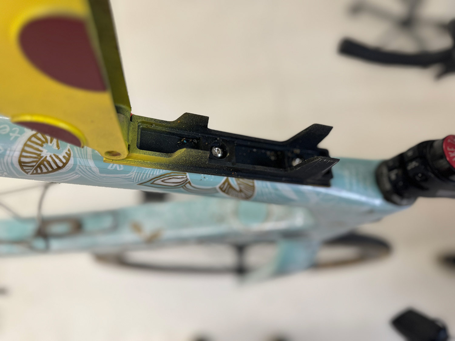Close-up of a colorful bicycle frame with a focus on the mounting area for accessories, showing a yellow and red attachment component and intricate design patterns.